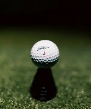 A Titleist golf ball on a black tee with a blurred green, inside Silverwood’s indoor golf lounge.