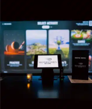 A touchscreen kiosk and drink menu rest on a table near a TrackMan golf simulator at the Kanata lounge.