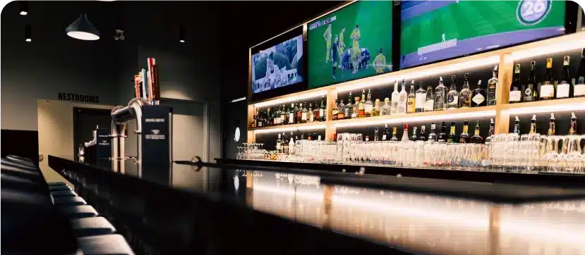 Long bar counter with backlit liquor shelves and wall-mounted screens showing golf at Silverwood’s Lounge.